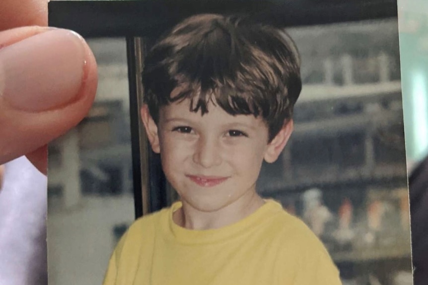 A photograph of a young boy in a yellow shirt and bowl hair cut smiling.