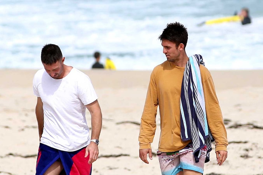 Two men walk together along a beach.