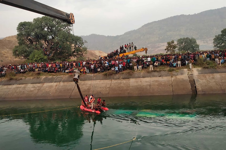 Rescuers in a dinghy look at a submerged bus being lifted out of a canal by a crane.