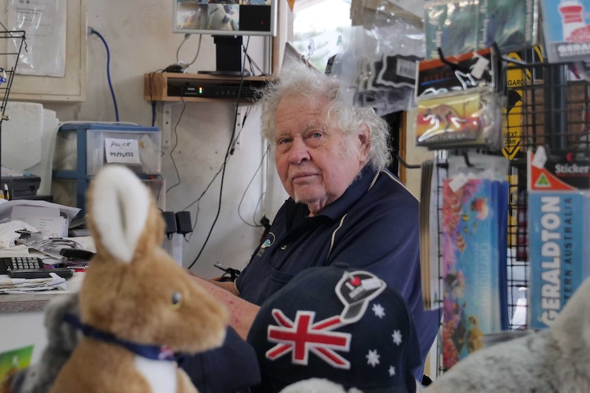 Gary Burns, an elderly gentlemen sits looking at the camera surrounded by souvenirs: magnets, soft toys, keyrings