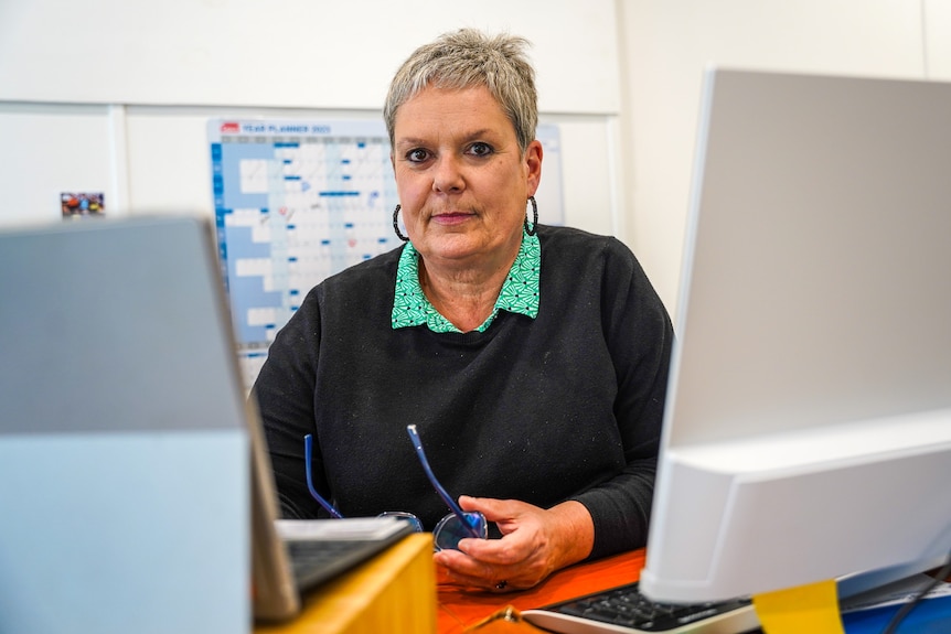 A woman with short grey hair wearing a jumper between two computer screens