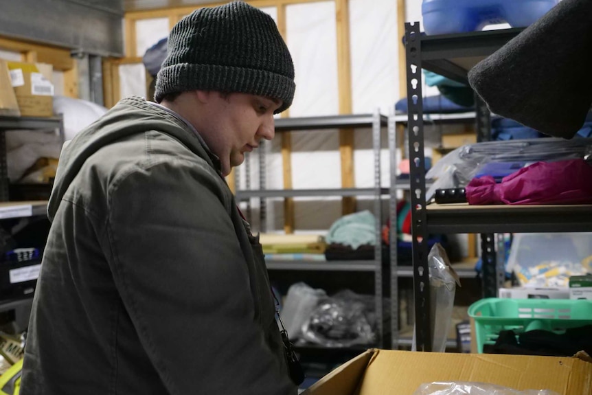A man wearing a coat and beanie in a storeroom with blankets and clothing on shelves