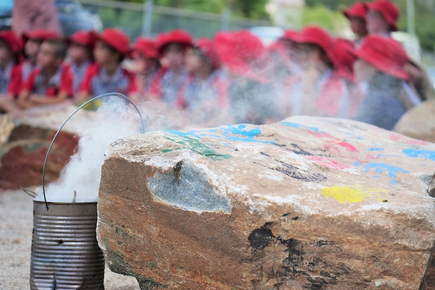 Multi-coloured hand prints on boulder, smoke coming out of tin, students in red and grey uniforms in background.