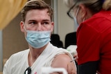 A young man wears a mask as he receives a vaccine from a nurse.
