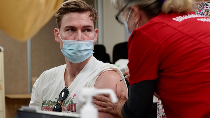 A young man wears a mask as he receives a vaccine from a nurse.