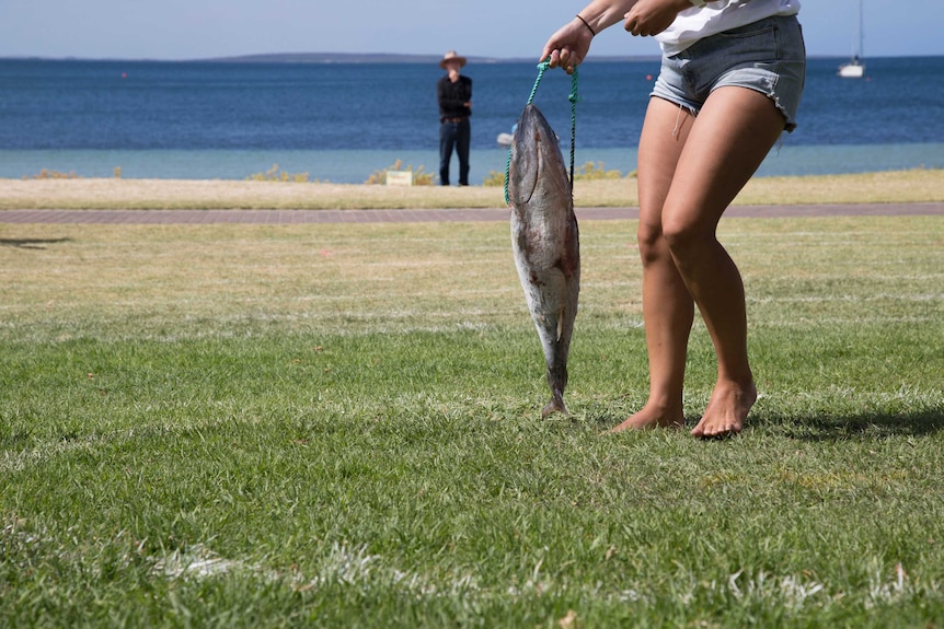A woman's legs seen from the waist down holds a tuna on a rope that is almost the same length as her legs, by the seaside.