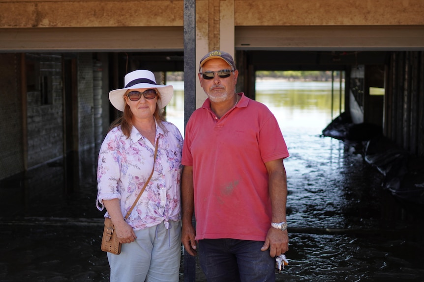 A man and a woman wearing hats and sunglasses standing in front of a house with the ground flooded