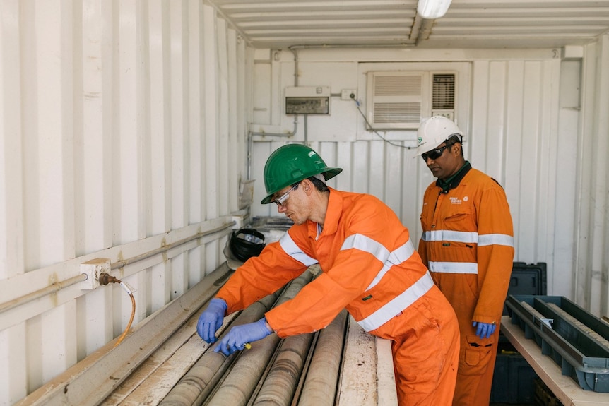 Two men wearing orange overalls look at pipes.