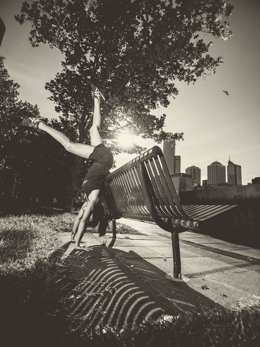 Black and white photo of Amy Han doing a handstand against a park bench with the sun and a tree in the background