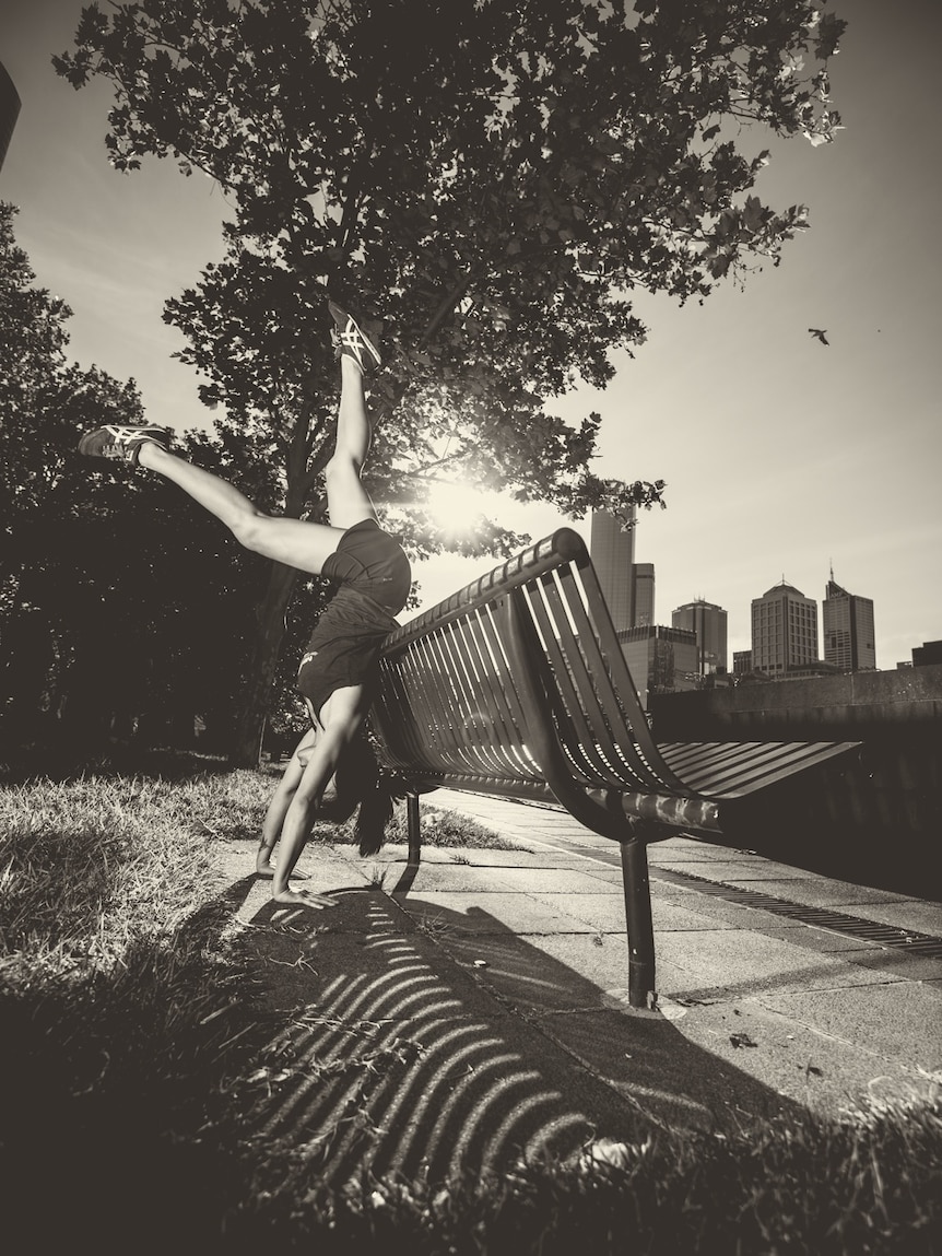 Photo en noir et blanc d'Amy Han faisant le poirier contre un banc de parc avec le soleil et un arbre en arrière-plan