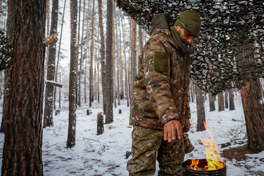 A soldier stands among snowy trees warming his hands over a fire