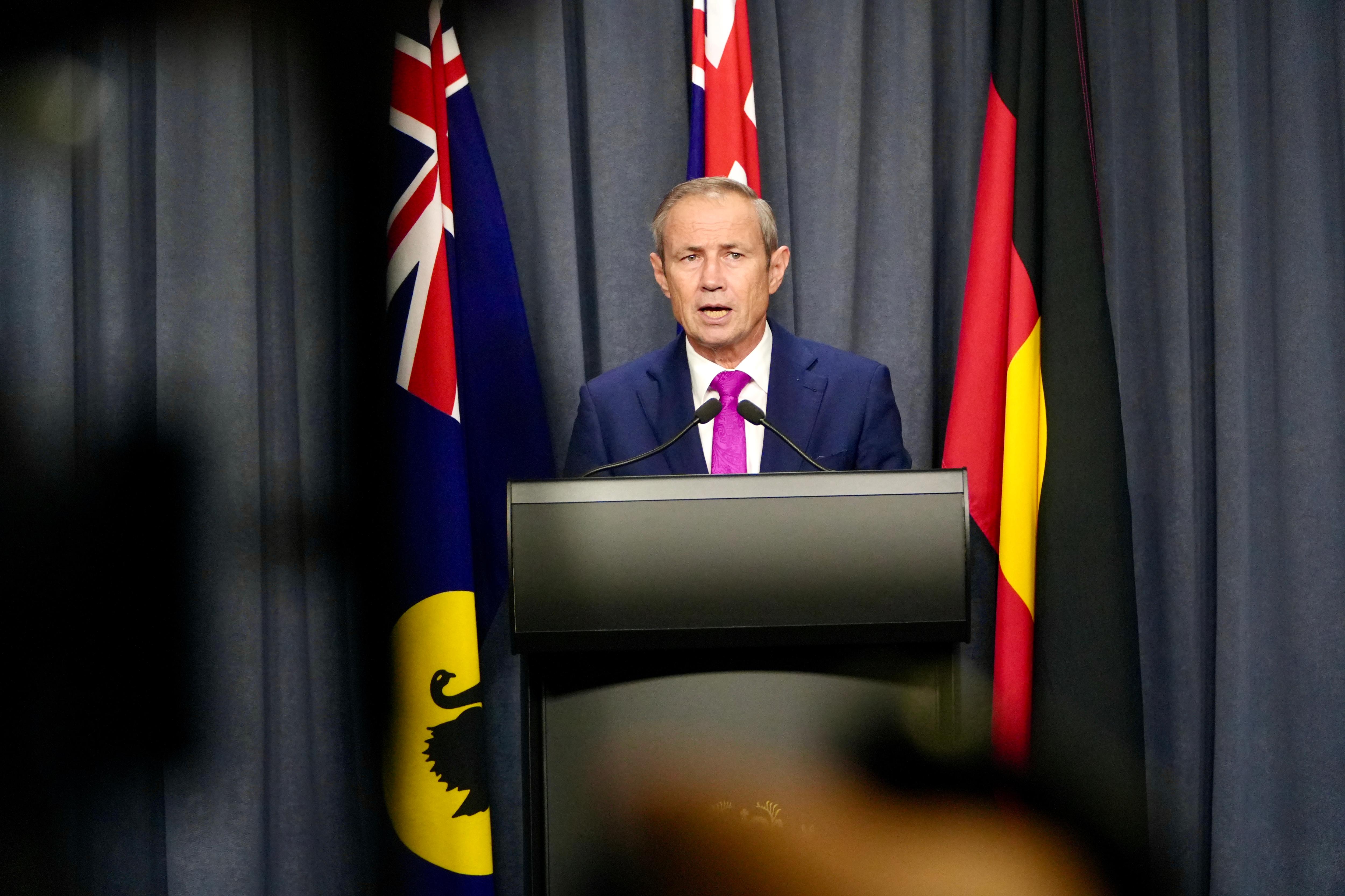 A wide shot of WA Premier Roger Cook speaking at a media conference indoors, wearing a suit and tie, flags behind him..