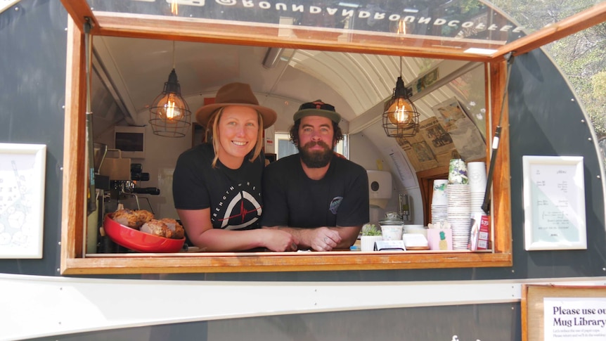 A man and woman smiling, looking out the window of a vintage-looking caravan that sells coffee.