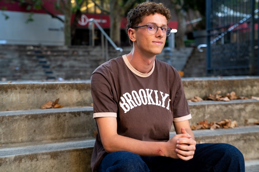 Young man with curly hair and glasses wearing brown t-shirt that reads 'Brooklyn', sitting on steps.