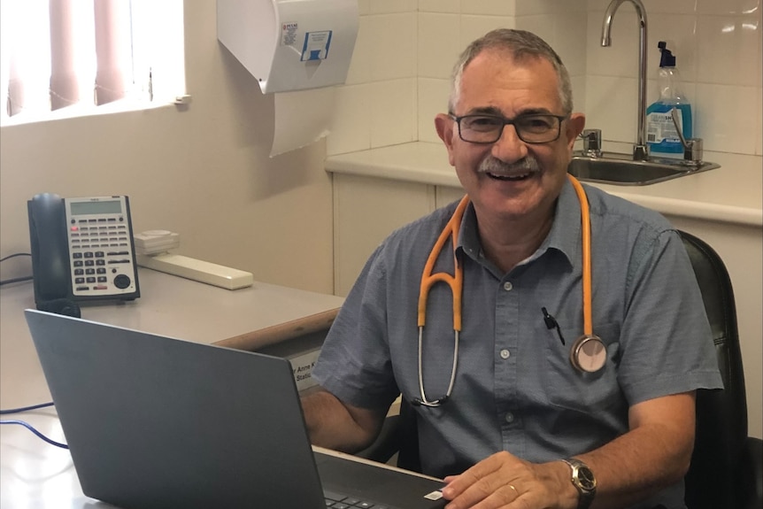 A smiling doctor with glasses and a mustache sits in his office.