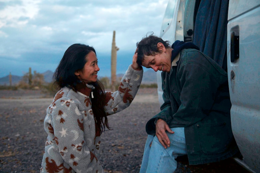 A woman with her hand in the hair of another woman sitting in the door of a van. 