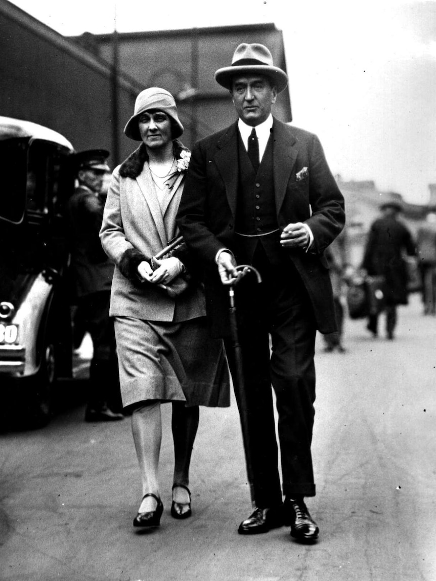 A black and white photo of a man in a suit and top hat walking alongside a smartly dressed woman