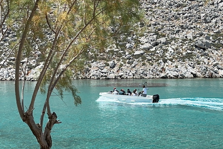 A motorboat carrying half a dozen people glides across clear blue water against a rocky mountainscape
