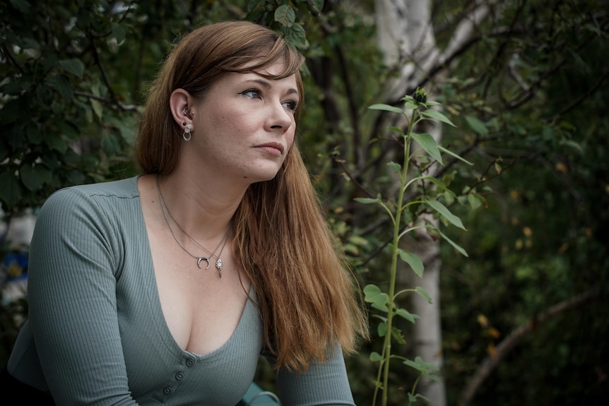 A woman in her 20s looks out into the distance, with trees and plants in the background.,