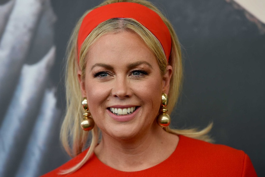 Samantha Armytage looks slightly to the left as she smiles. She wears a red dress, matching headband and gold earrings.