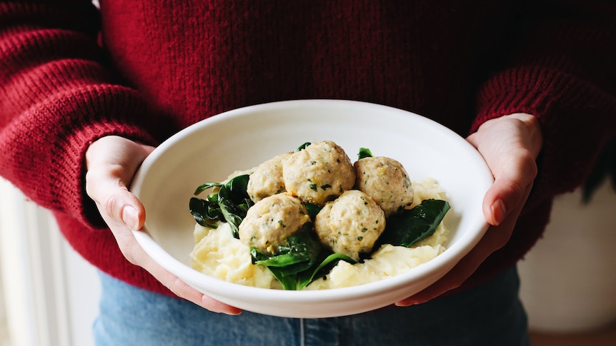 A person holds a bowl containing chicken meatballs sitting on top of mashed potato and greens.