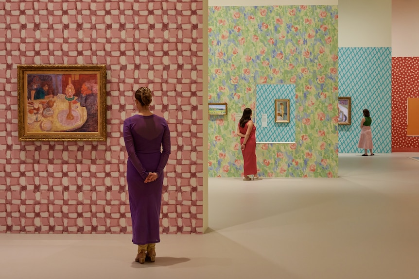 A woman looks at a painting on a bright patterned wall. In the background, two other women do the same.