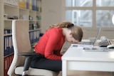 A woman wearing a red jumped sleeps with her head on a desk in front of an open laptop.