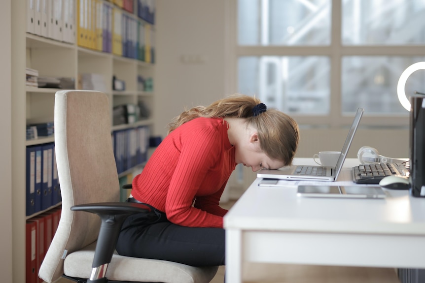 A woman wearing a red jumped sleeps with her head on a desk in front of an open laptop.
