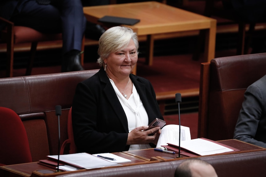 McMahon is sitting at her senate desk, looking up, smiling, holding phone in right hand.