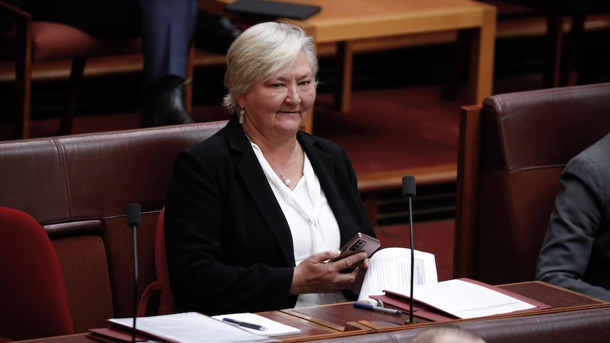 McMahon is sitting at her senate desk, looking up, smiling, holding phone in right hand.