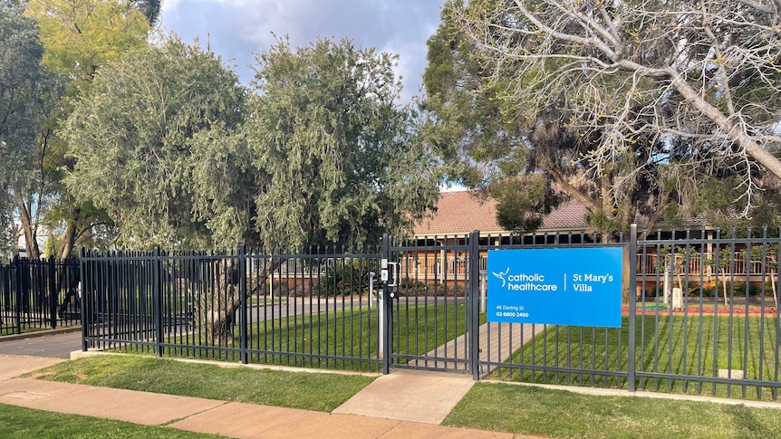 A black fence with a blue sign on it surrounds a building with trees.