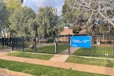 A black fence with a blue sign on it surrounds a building with trees.