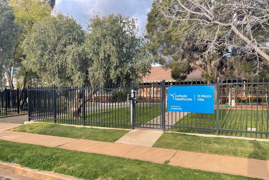 A black fence with a blue sign on it surrounds a building with trees.
