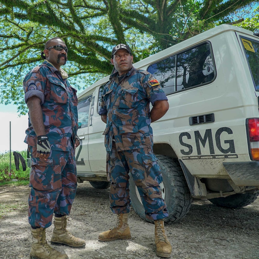 Two PNG men in camouflage uniforms stand under a lush green tree
