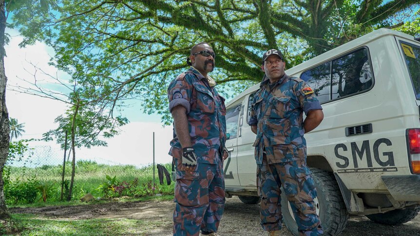 Two PNG men in camouflage uniforms stand under a lush green tree