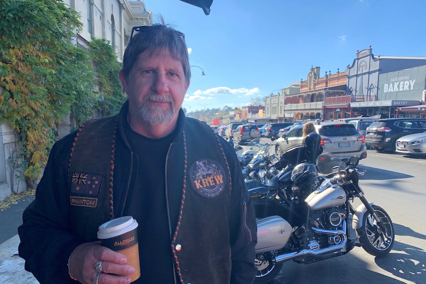 A man wearing motorcycle gear poses for a photograph while standing on a small town's main street while holding a coffee.