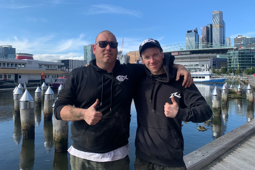 Two men stand on a river boardwalk in front of the city.