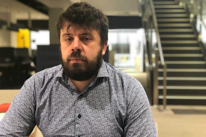 David Gardner, looking serious, faces the camera. A set of stairs is in the background.