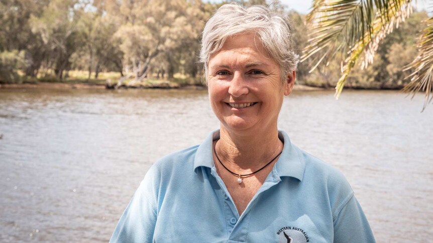 A woman with short grey hair, wearing a pale blue polo shirt, smiles as she stands in front of a body of water.