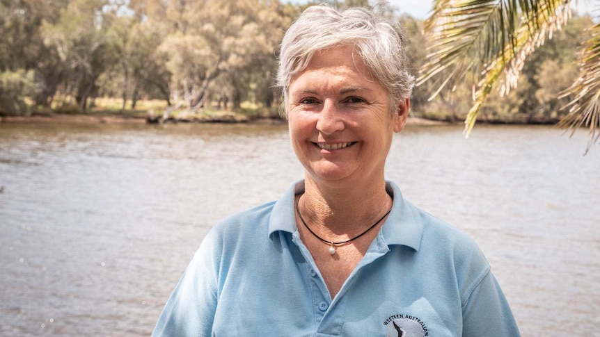 A woman with short grey hair, wearing a pale blue polo shirt, smiles as she stands in front of a body of water.