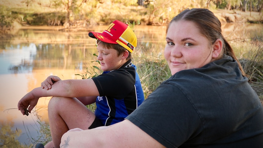 Jordy sits on the bank of a river in rural Queensland, with a friend wearing a colourful cap.