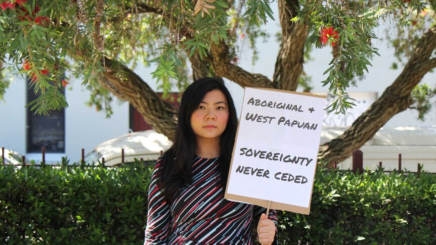 A woman standing under a tree holds a sign reading, "Aboriginal and West Papuan sovereignty never ceded"