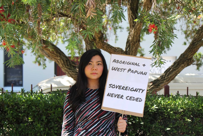 A woman standing under a tree holds a sign reading, "Aboriginal and West Papuan sovereignty never ceded"