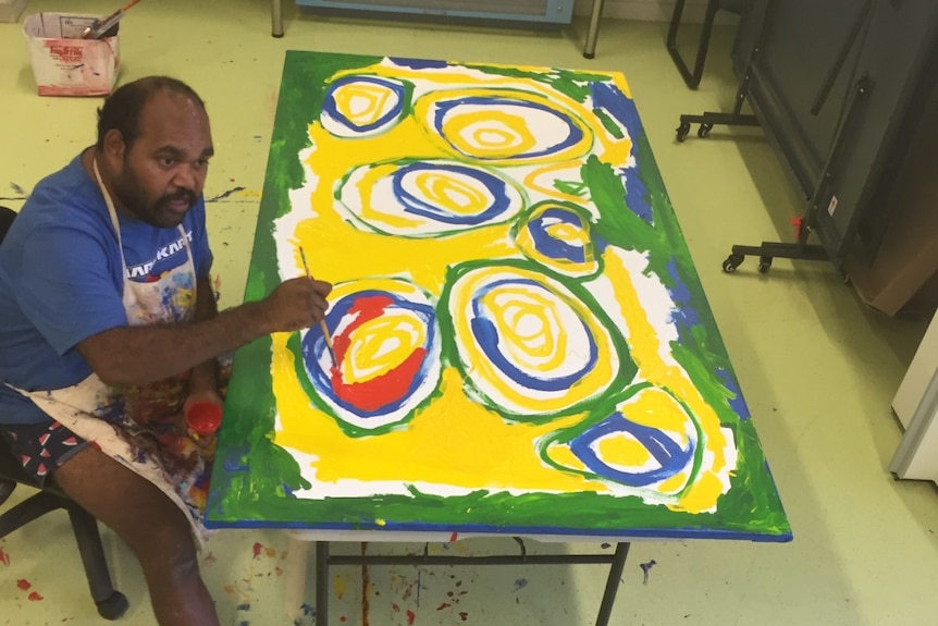 Indigenous man wearing a blue top, shorts and white apron sits next to a large canvas on a table painting.