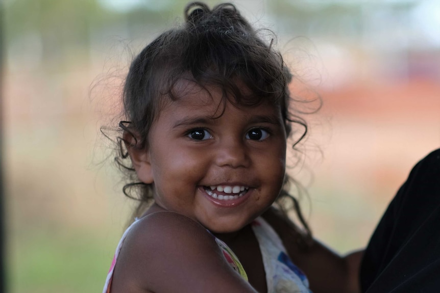 A young Indigenous girl with curly hair and bright eyes smiles widely for the camera.