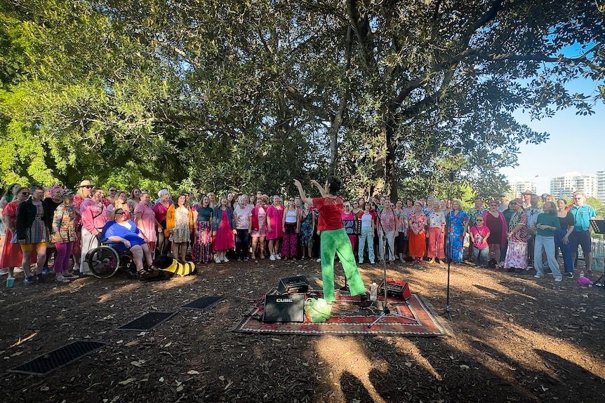 A woman conducts a choir or brightly coloured performers, outside under a tree