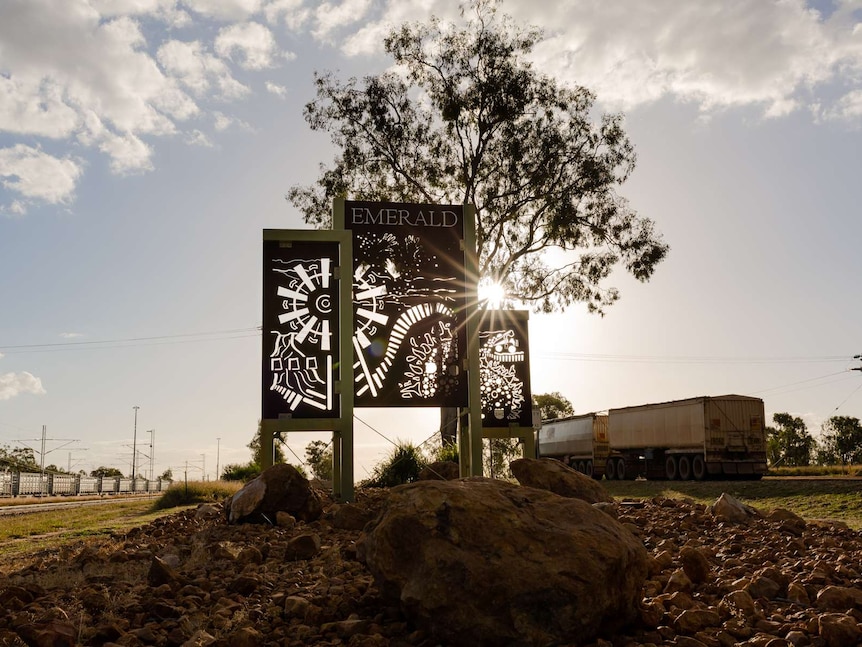 A metal sign with carved out rail/windmill/stones reads 'Emerald'. Semi-trailer passing in background and cattle train visible.