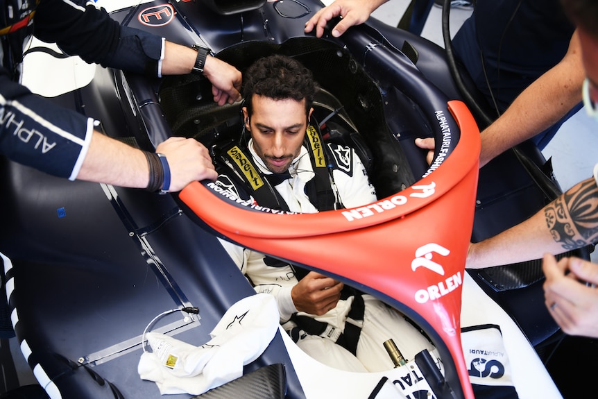 An F1 driver, sitting in his car, no helmet on, as the hands of mechanics are seen making adjustments.