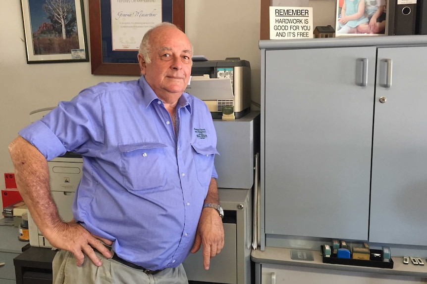 A man stands in an office next to cupboards wearing a blue shirt.