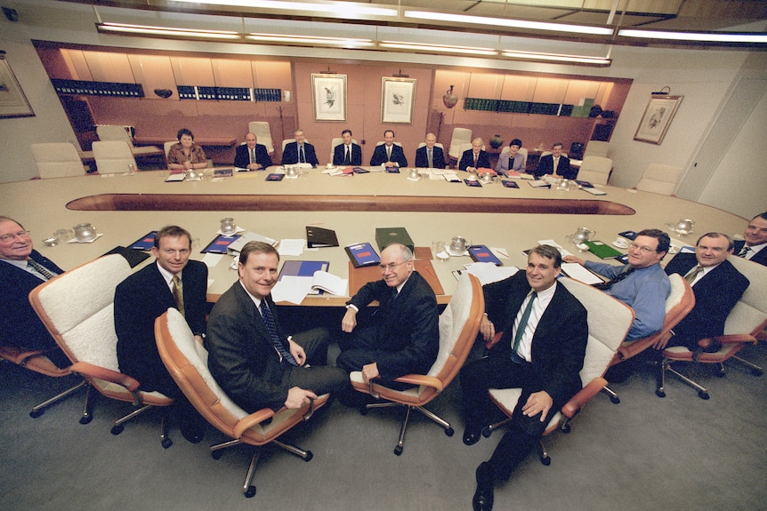 A wide shot of the cabinet room mostly men, sitting around a large table. John Howard is in the middle on the closest side